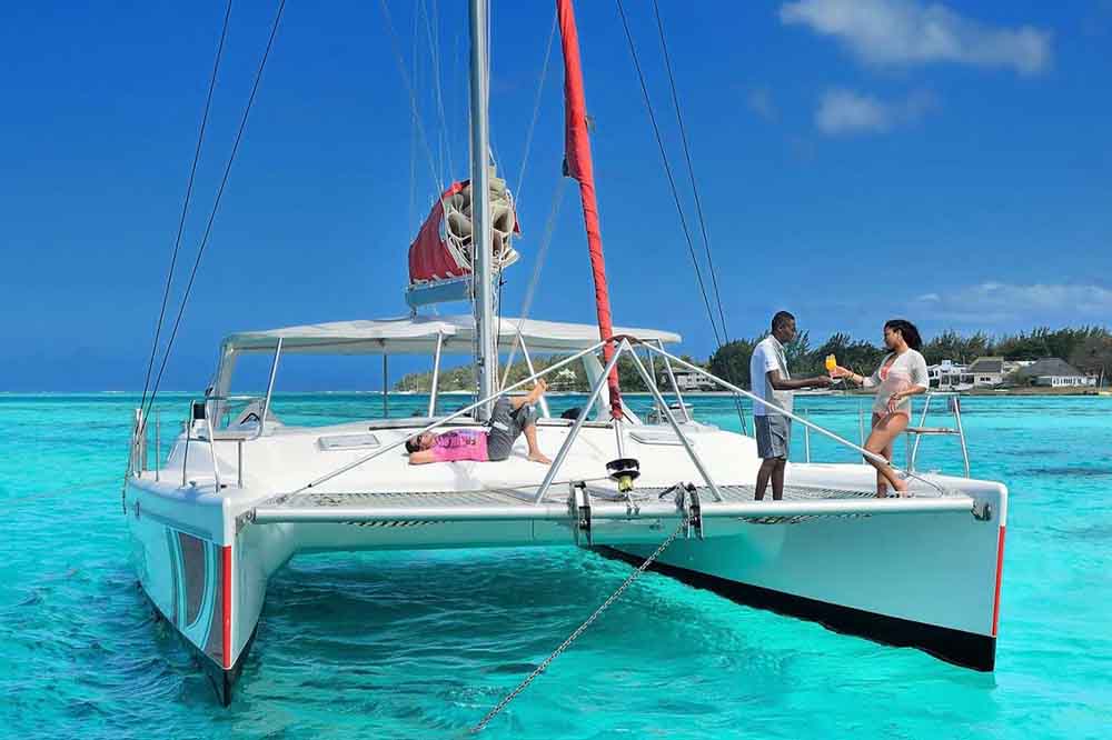 A white catamaran sails on vibrant turquoise water under clear blue skies. A person relaxes on the deck, while two people stand at the edge, sharing a drink.
