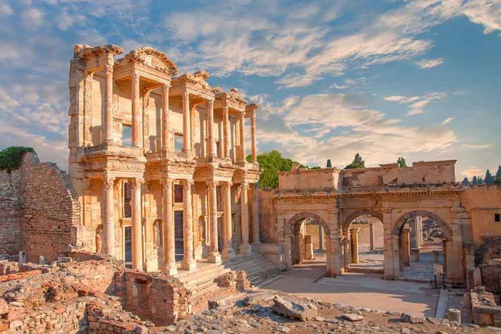 Ancient ruins of a grand, two-story stone library and adjacent arches under a partly cloudy sky, conveying historical grandeur and timelessness.