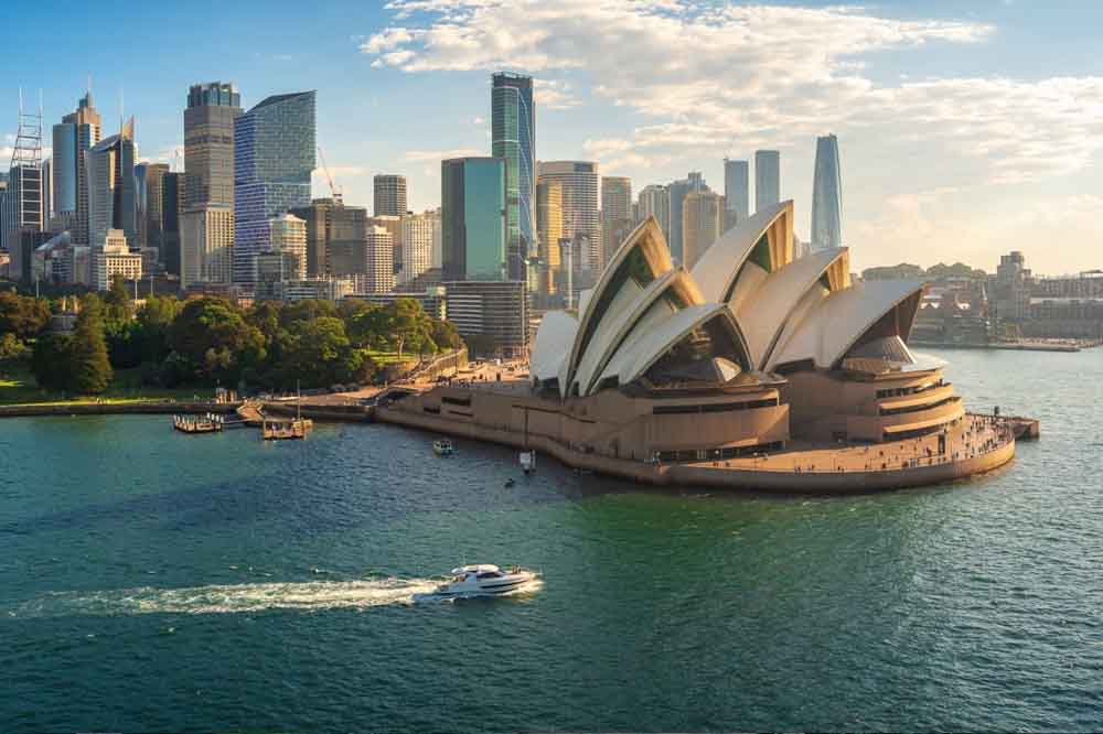 Aerial view of the Sydney Opera House with its iconic sails by the waterfront. Skyscrapers and lush greenery form the background. A boat cruises nearby.