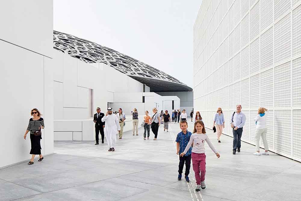 People walk on a modern architectural pathway, featuring white walls and a lattice roof. The scene is bright and lively, with a clear sky overhead.