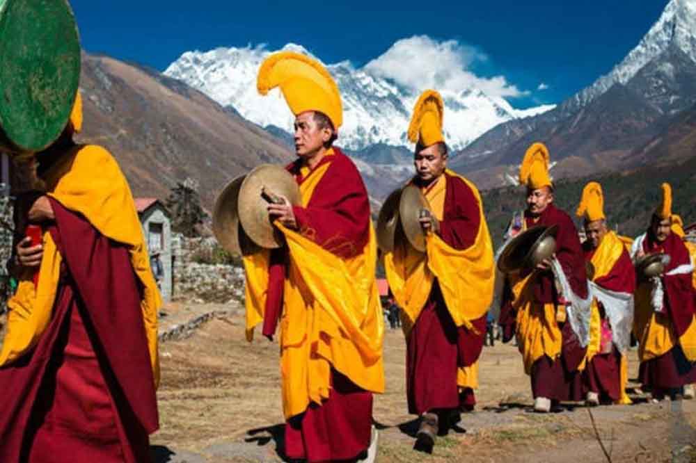 Monks in orange and maroon robes play cymbals, walking in a line against a backdrop of snow-capped mountains under a clear blue sky.