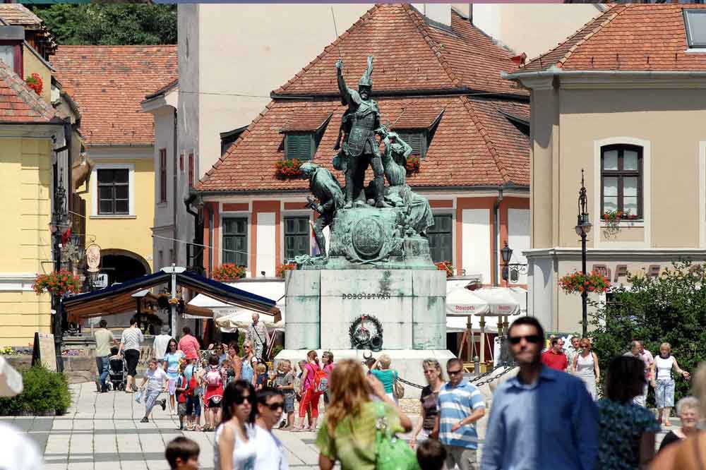 Bustling town square with a large statue at the center. People stroll around under clear skies, surrounded by charming, historic buildings.