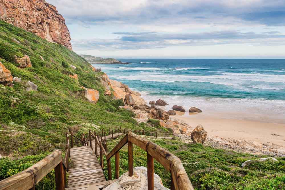 Wooden stairs lead down a lush, green cliffside to a sandy beach with rocky edges. The ocean waves gently crash under a cloudy sky, conveying tranquility.