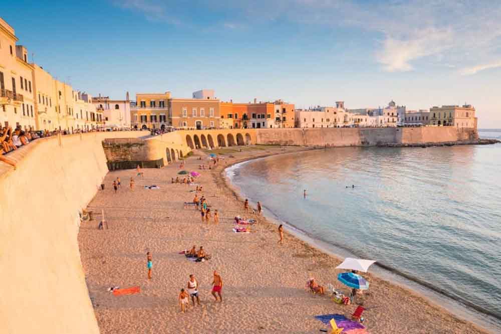 Serene beach scene at sunset with relaxed people on sandy shore; colorful buildings and a historic wall line the backdrop, reflecting a peaceful ambiance