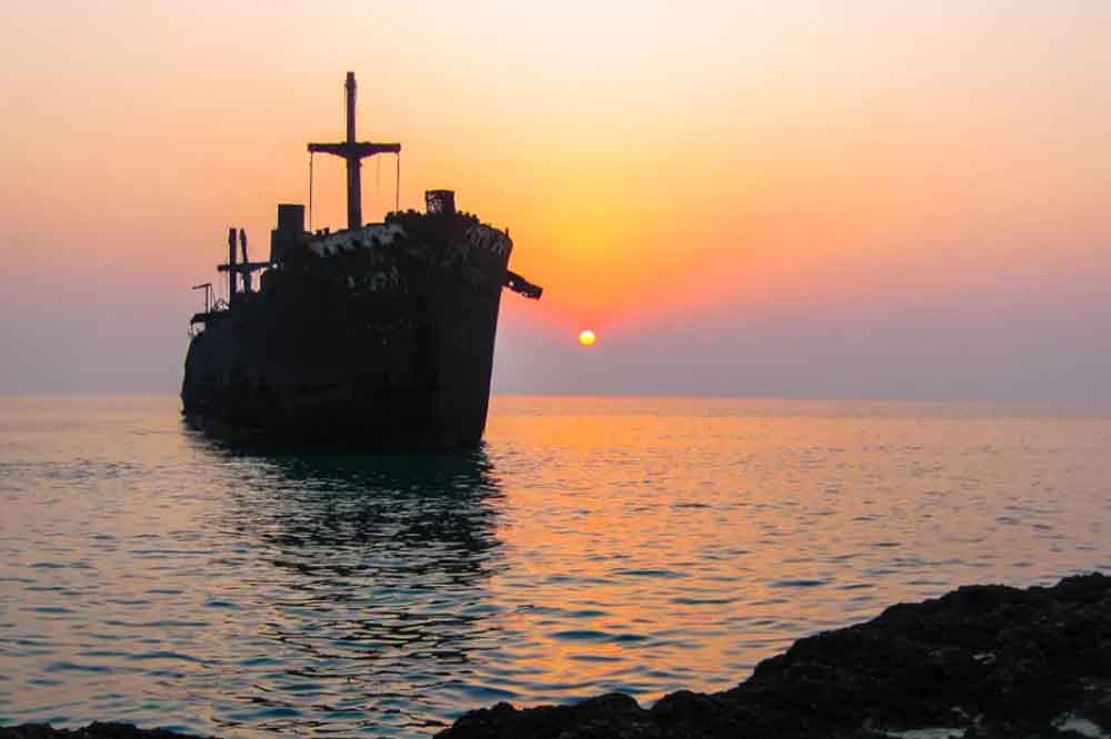 Silhouette of an abandoned ship at sea against a vibrant sunset, with the sun setting on the horizon. Calm waters reflect the orange and pink sky.