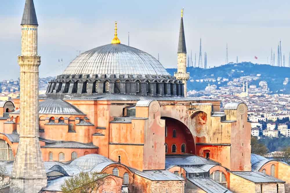 Historic building with a large central dome, flanked by two minarets. The structure is reddish with intricate details; cityscape in the background. Located in Turkey. 