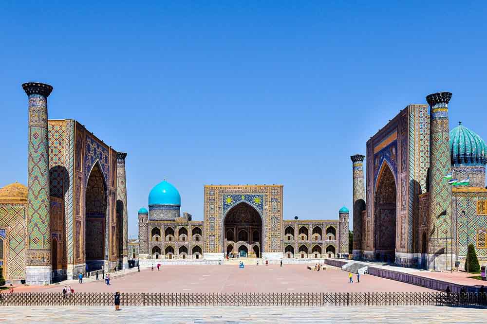 Spacious courtyard in front of majestic, intricate buildings with vibrant blue domes under a clear sky. Few people walking, conveying a serene atmosphere.
