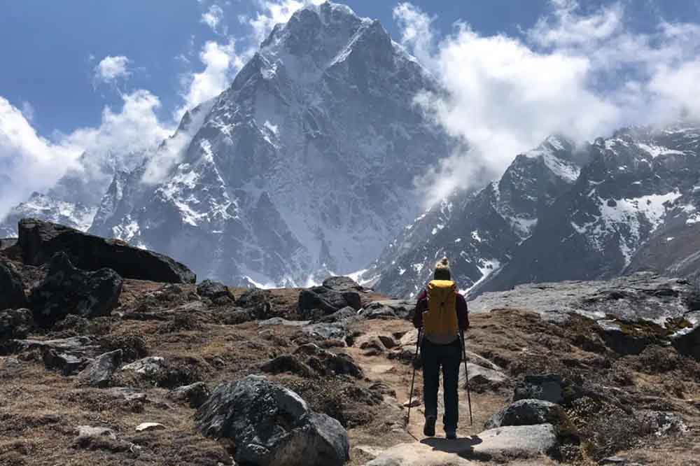 Hiker with yellow backpack and walking sticks stands on a rocky path, gazing at a snowy mountain peak under a partly cloudy blue sky. Majestic and serene.