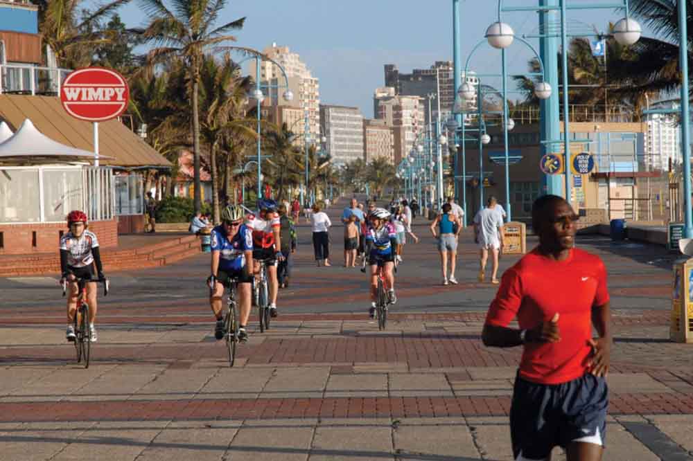 Cyclists and a jogger on a sunny boardwalk lined with palm trees and lampposts. A 