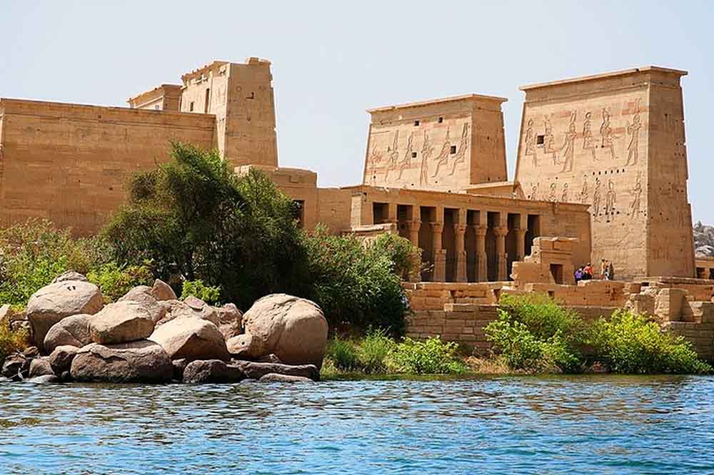 Ancient Egyptian temple by a river, featuring sandy stone walls adorned with hieroglyphics and columns. Lush greenery and rocks in the foreground.