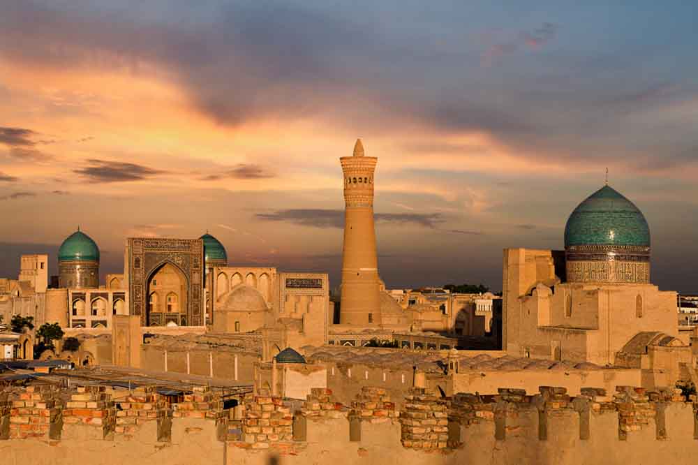 Ancient cityscape at sunset with a tall minaret and blue-domed structures contrasting against a vibrant orange sky. The scene exudes historical grandeur.