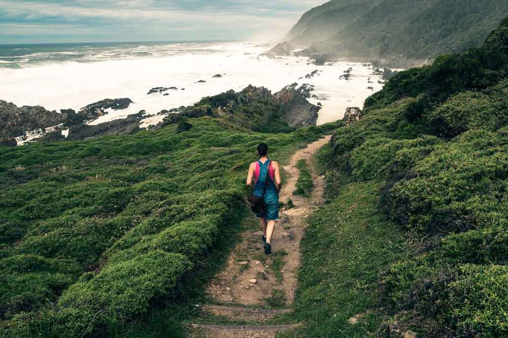 A person in athletic wear hikes along a lush, green coastal trail. Ocean waves crash on rocky cliffs under a cloudy, moody sky, evoking a sense of adventure.