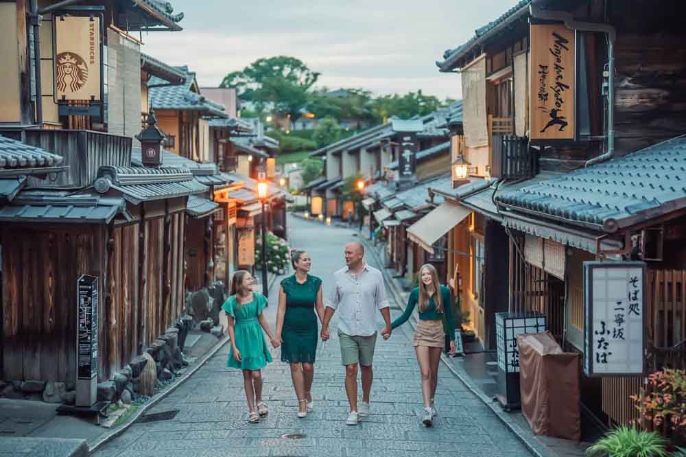 A family of four, holding hands, strolls down a charming, traditional Japanese street at dusk, surrounded by historic wooden buildings and warm lanterns.