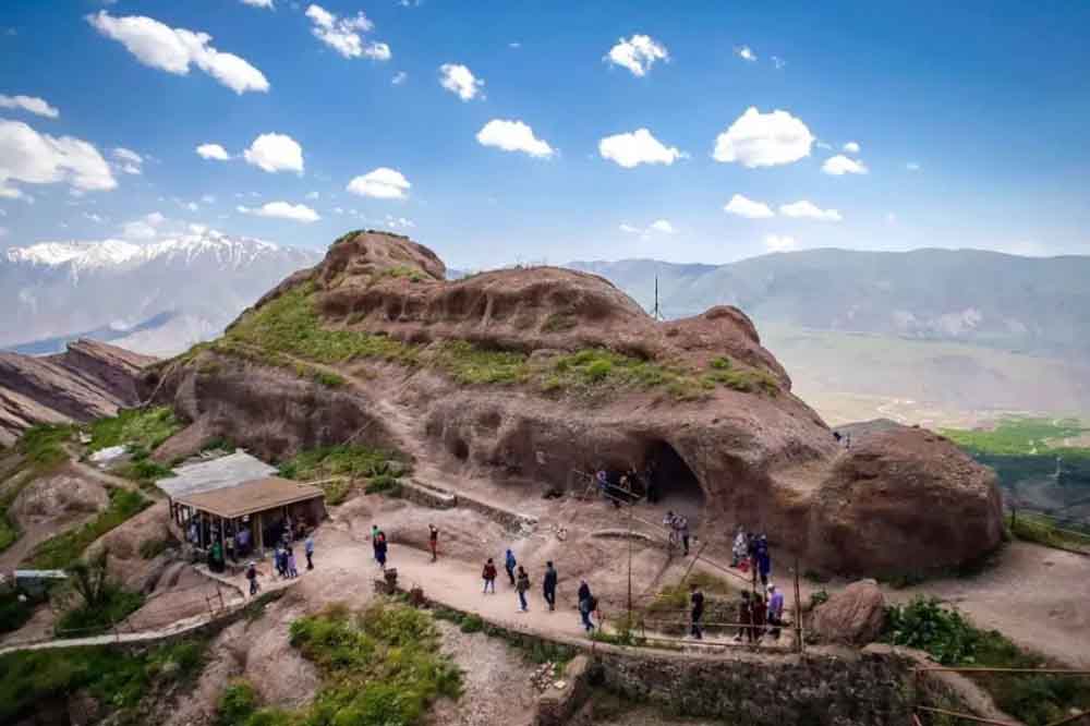 Rocky landscape featuring a natural cave formation with people exploring. Lush greenery covers parts of the rock. Snow-capped mountains and blue sky in the background.