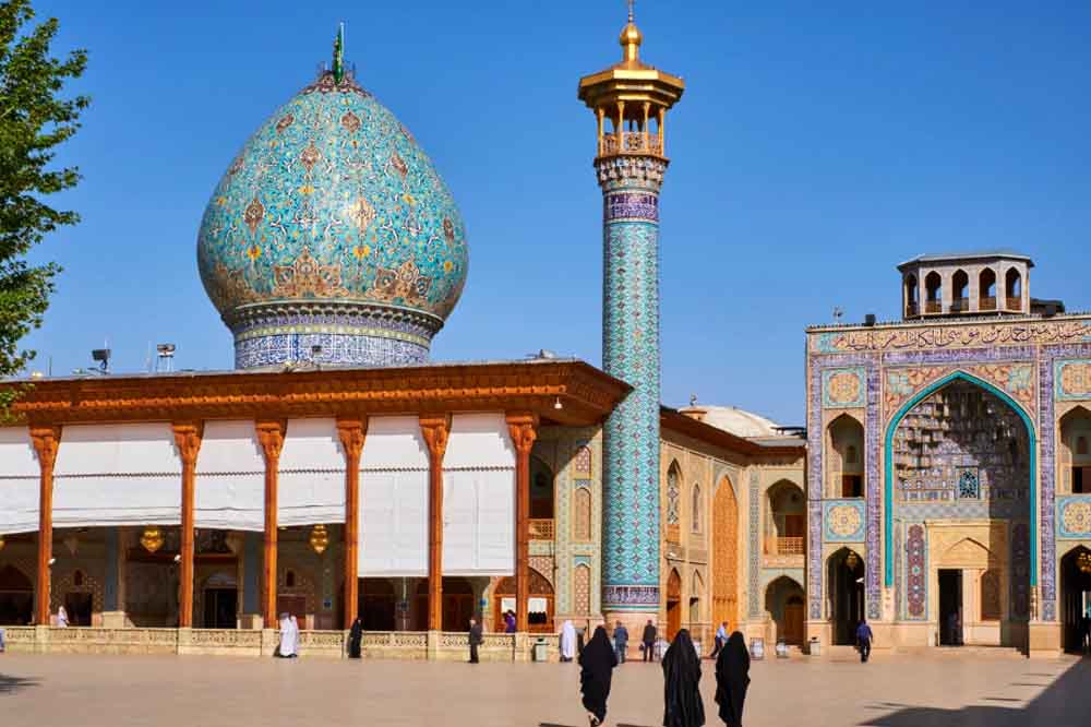 Islamic architecture featuring a large, intricate turquoise dome and a slender minaret. People walk in a sunlit courtyard with arched entrances.