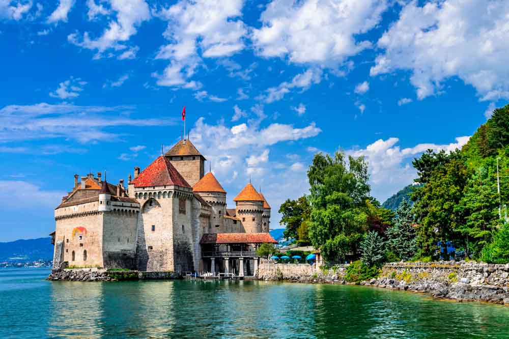 Medieval stone castle with conical roofs on a serene lake, surrounded by lush greenery and a bright blue sky with scattered clouds. Tranquil and picturesque.
