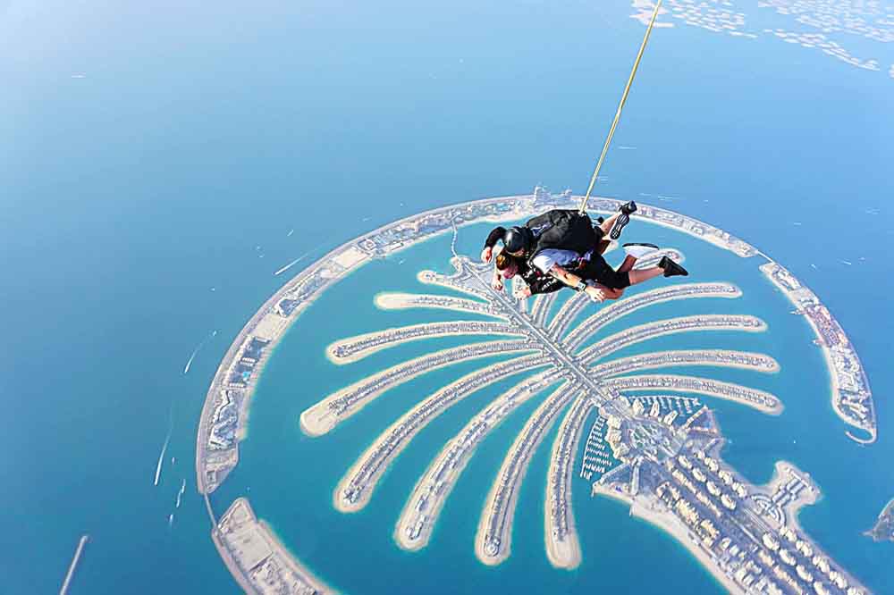 Two skydivers in tandem freefall above Palm Jumeirah, Dubai. The island's palm shape is prominent against the blue sea, evoking excitement and adventure.