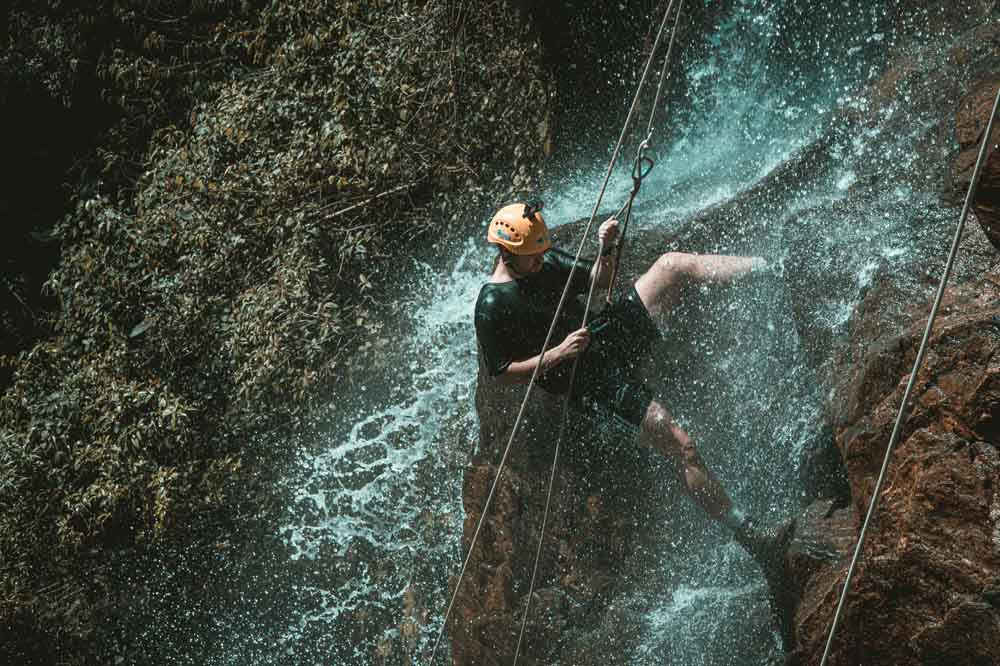 Person in a helmet rappelling down a waterfall on a rocky cliff, surrounded by splashing water. An adventurous and thrilling scene.