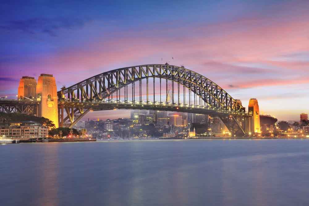 Sydney Harbour Bridge at dusk, illuminated with warm lights against a vibrant pink and blue sky. The city skyline glows softly in the background.