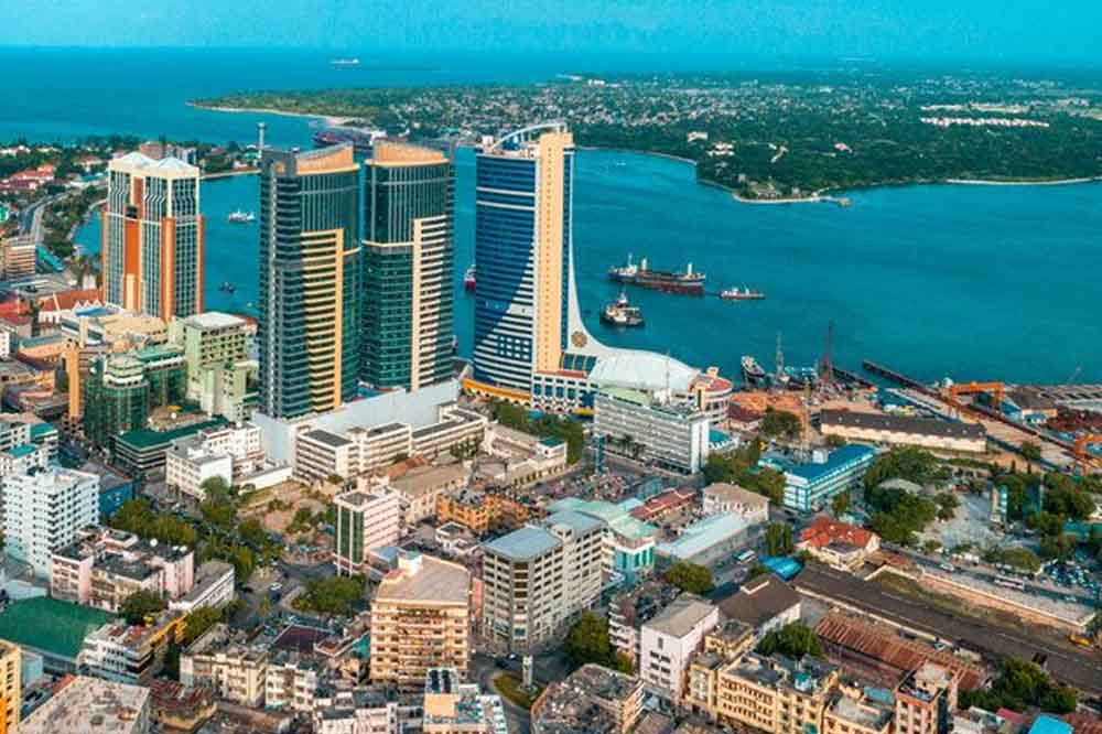 Aerial view of a cityscape with tall modern skyscrapers near a calm blue bay. Boats are visible on the water, and urban buildings surround the area.