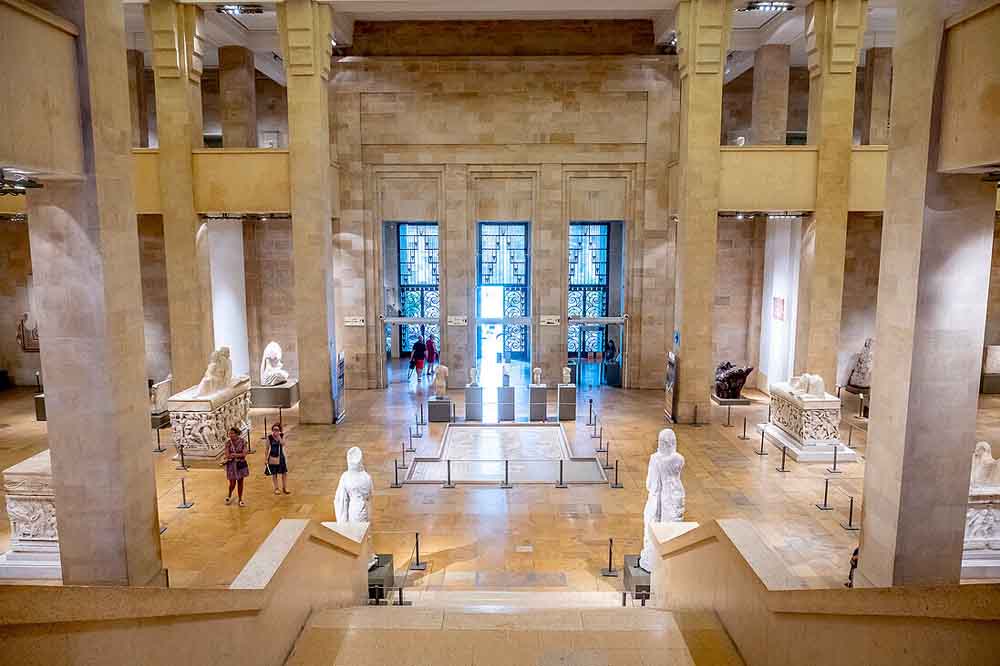 Expansive museum hall with tall beige columns, classical sculptures, and a checkered floor. Two visitors observe art, invoking a serene, reflective mood.