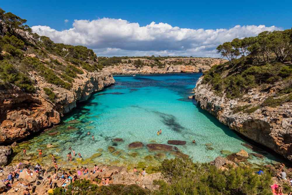 A scenic coastal inlet with clear turquoise water surrounded by rocky cliffs and lush greenery. People are swimming and sunbathing, exuding a relaxed, joyful vibe.