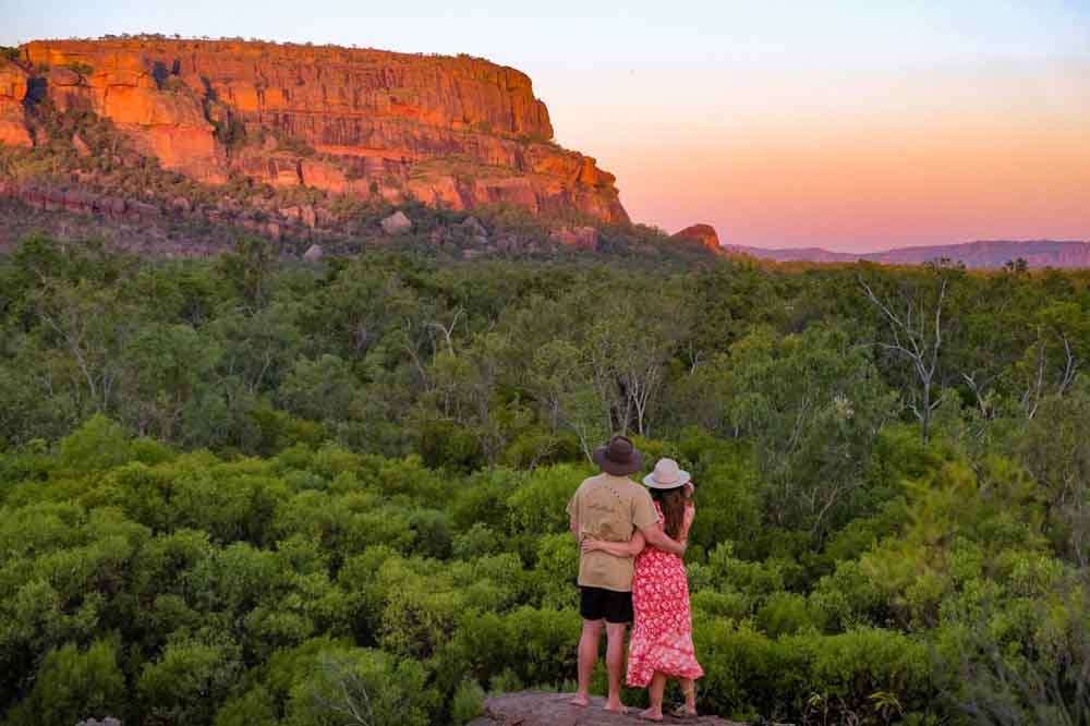 A couple stands embracing, gazing at a sunlit, reddish cliff surrounded by dense greenery under a colorful sunset sky, conveying tranquility and awe.